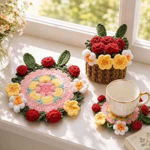 May include: A set of three crocheted items including a large circular floral mat, a smaller circular coaster holding a white ceramic cup with a gold rim, and a brown crocheted basket filled with red, yellow, and white crocheted flowers.