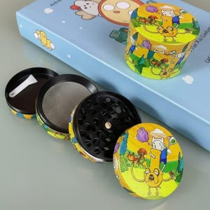 May include: A four-piece herb grinder with a cartoon design featuring characters in a yellow and green landscape. The grinder is disassembled, showing the different components, including a mesh screen and a small scoop.