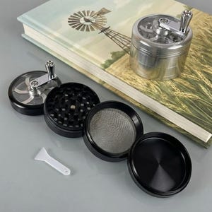 May include: A disassembled silver and black herb grinder with a crank handle. The grinder is shown with its various components: a top with a clear window, a grinding chamber, a mesh screen, and a bottom chamber. A small white scraper is also included.