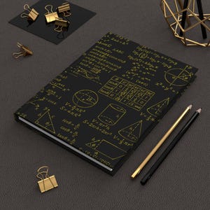 May include: A black notebook adorned with gold mathematical formulas and diagrams. Gold binder clips, pencils, and a geometric desk organiser surround the journal. The cover displays equations and geometric shapes.
