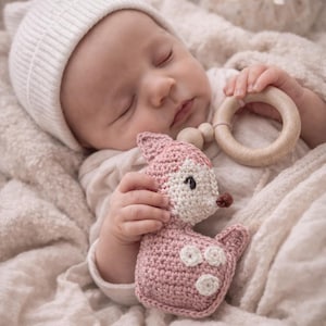 Puede incluir: Un bebé dormido con gorro de punto blanco y body, acurrucado en una manta suave. El bebé sostiene un juguete de ciervo rosa tejido a ganchillo y un anillo de dentición de madera. El ciervo tiene detalles blancos y una nariz marrón.