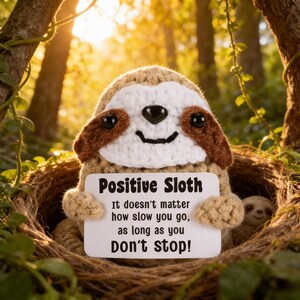 May include: A crocheted sloth toy with a white face, brown accents, and black eyes, holding a sign that reads "Positive Sloth." The sign also includes the text "It doesn't matter how slow you go, as long as you Don't stop!" Another sloth is visible in the background.