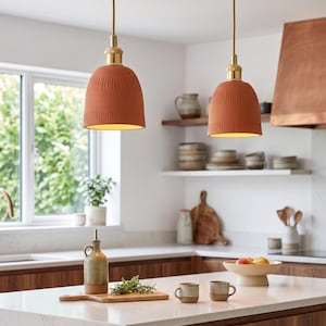 Pode incluir: Duas luminárias pendentes de terracota com acessórios de latão pendem sobre uma ilha de cozinha. As luzes têm uma superfície texturizada e emitem um brilho quente. A cozinha apresenta armários de madeira, uma bancada branca e uma coifa de cobre.