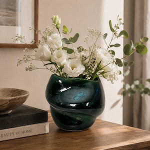 Peut inclure: Un vase en verre vert foncé avec un design torsadé unique, rempli de fleurs blanches et de verdure. Le vase est posé sur une surface en bois, à côté d'une pile de livres et d'un bol décoratif. Les fleurs sont des roses blanches et du gypsophile.