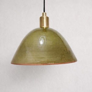 Op de afbeelding: Een groene keramische hanglamp met een messing fitting. De lamp heeft een afgeronde, klokvormige kap met een glanzende afwerking. De kap heeft een gedempte groene kleur en de fitting hangt aan een zwart snoer.