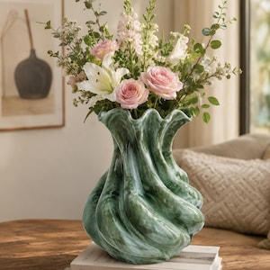 Peut inclure: Un vase vert texturé avec un design tourbillonnant unique, rempli d'un bouquet de roses roses, de lys blancs et d'autres fleurs. Le vase est posé sur une pile de livres, dont l'un porte l'inscription "LIVE BEAUTIFUL."