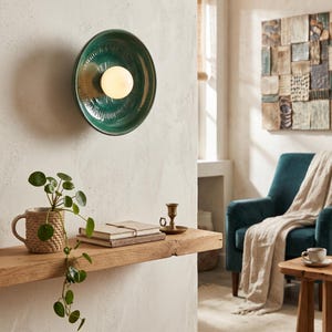 Può includere: Una lampada da parete circolare composta da una ciotola in ceramica smaltata verde scuro con linee strutturate radianti e una lampadina sferica in vetro bianco al centro. Il design crea un bagliore focalizzato sulla superficie della parete.