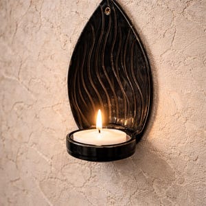 Peut inclure: Un bougeoir mural en métal noir en forme de flamme, contenant une bougie blanche allumée. Le bougeoir présente un motif de vagues décoratives et est fixé à un mur beige texturé. La flamme de la bougie est vive.
