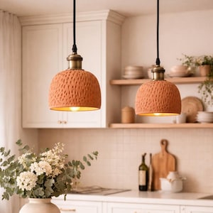 Peut inclure: Deux suspensions en terre cuite de couleur terracotta avec des abat-jours texturés sont suspendues à des cordons noirs. Les luminaires ont des fixations en laiton et sont suspendus au-dessus d'un plan de travail blanc. La cuisine comprend un vase de fleurs et des étagères avec de la vaisselle.