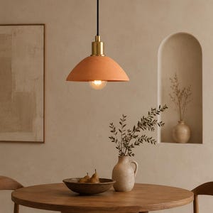 Pode incluir: Uma luminária pendente com uma cúpula fosca na cor pêssego, suspensa por um fio preto em um suporte de tom latão. A luz inclui uma lâmpada exposta que emite um brilho quente sobre uma mesa redonda de madeira com uma tigela de peras.