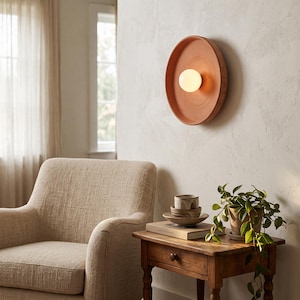 May include: A circular terracotta wall sconce featuring a central, glowing white spherical light bulb. The fixture has a smooth, matte finish and a shallow, bowl-like shape that extends from the wall, providing warm illumination.