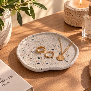 Op de afbeelding: Een ronde, gebroken witte keramische schaal met een veelkleurig gespikkeld patroon in blauw, roze en zwart. Twee goudkleurige metalen ringen en een goudkleurige ketting met een ronde hanger liggen op het oppervlak van de schaal.