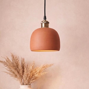 Peut inclure: Suspension luminaire en terre cuite de couleur terracotta, avec un abat-jour arrondi, suspendu par un câble noir. Le raccord est de couleur laiton. Un vase avec des herbes de la pampa séchées est en dessous, sur fond rose.