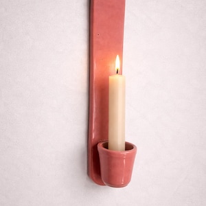 Peut inclure: Un bougeoir mural en céramique rose avec une bougie blanche allumée. Le support a une longue plaque arrière rectangulaire et une petite coupelle pour tenir la bougie. La bougie est de couleur ivoire et a une flamme visible.
