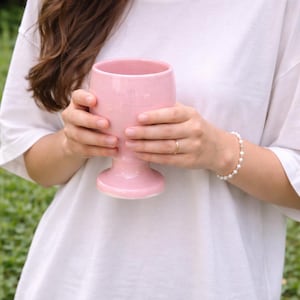 Peut inclure: Un gobelet en céramique rose clair avec un large bol et une base en piédestal. Le gobelet est tenu par une personne portant un t-shirt blanc. L'arrière-plan est un feuillage vert flou.