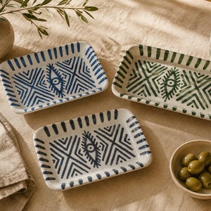 Peut inclure: Trois plateaux rectangulaires en céramique aux coins arrondis, ornés de motifs géométriques peints à la main et d'un motif central en forme d'œil. Un plateau présente des motifs bleus, tandis que les deux autres sont vert foncé et noirs.