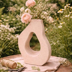 Peut inclure: Un vase beige de forme abstraite avec une grande ouverture, rempli de roses roses et de petites fleurs blanches. Le vase est posé sur une table en bois avec des ciseaux, de la ficelle et des feuilles, dans un jardin.