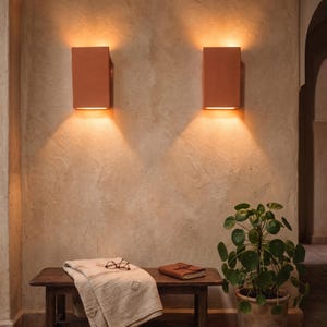 May include: Two rectangular, terracotta-colored wall sconces emit warm light, casting a glow on the textured beige wall. A wooden bench holds a folded blanket, glasses, and a leather-bound journal. A potted plant with round leaves sits nearby.