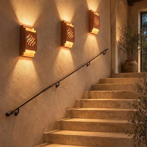 May include: Warm-toned outdoor lighting fixtures with a wave-like design are mounted on a textured wall. A staircase with a black handrail leads upwards. The scene is illuminated by the fixtures, creating a welcoming ambiance.