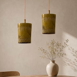 Puede incluir: Dos lámparas colgantes cilíndricas con un esmalte verde oliva brillante y textura acanalada vertical, suspendidas por varillas metálicas delgadas. Las lámparas presentan un acabado liso en la parte superior e inferior, con un color uniforme.