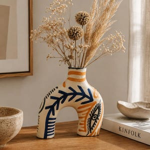Op de afbeelding: Een boogvormige keramische vaas met een crèmekleurige basis, versierd met handgeschilderde marineblauwe bladpatronen, oranje strepen en een zwart oogmotief. De vaas bevat een droogbloemstuk met hoge grassen en kleine ronde zaadhoofdjes.