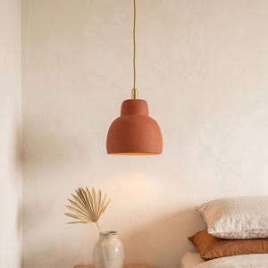 Könnte beinhalten: Eine terracottafarbene Pendelleuchte mit rundem Schirm hängt an einem goldfarbenen Kabel. Die Leuchte hängt über einem Nachttisch mit einer Vase und einem getrockneten Palmblatt. Die Schlafzimmereinrichtung umfasst ein Bett mit Kissen und Bettwäsche.