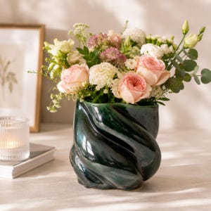 Peut inclure: Un vase en céramique vert foncé et brillant, au design torsadé, contient un bouquet de roses roses et blanches et d'autres fleurs. Le vase est posé sur une surface de couleur claire, avec un cadre photo et une bougie en arrière-plan.