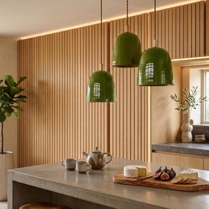 Peut inclure: Trois lampes suspendues vertes au-dessus d'un îlot de cuisine. Les lampes ont une forme de cloche et sont suspendues par des cordons marron. Une planche à découper en bois avec du fromage et des figues est posée sur l'îlot.