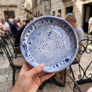 Peut inclure: Un bol rond en céramique avec une base bleu clair et un motif tacheté bleu foncé. Le bol présente une finition lisse et brillante ainsi qu'un bord légèrement surélevé, mettant en valeur une esthétique artisanale avec des éclaboussures.