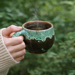 Può includere: Una tazza in ceramica fatta a mano con base marrone scuro e bordo verde turchese, piena di una bevanda calda. Il vapore sale dalla bevanda, suggerendo calore. La tazza è tenuta in mano su uno sfondo verde sfocato.