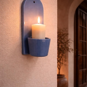 Peut inclure: Un bougeoir mural en céramique bleu avec une bougie ivoire allumée. Le bougeoir a un dessus incurvé et un petit support en forme de coupe pour la bougie. Le fond est un mur texturé de couleur claire.