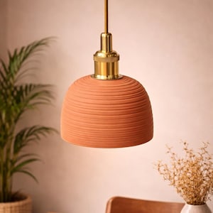 May include: A terracotta pendant light with a ribbed, bell-shaped shade. The light fixture has a brass-coloured socket and a brown cord. The light hangs above a wooden table, with a plant and vase in the background.