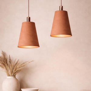 Puede incluir: Dos lámparas colgantes de color terracota con herrajes cobrizos cuelgan del techo. Las lámparas tienen forma cónica y emiten un brillo cálido. Un jarrón con plantas secas y un cuenco descansan sobre un mueble de madera.