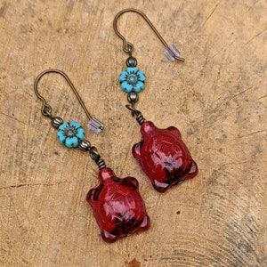 May include: A pair of earrings featuring a red glass turtle charm and a turquoise blue flower bead. The earrings are made with bronze-colored metal.