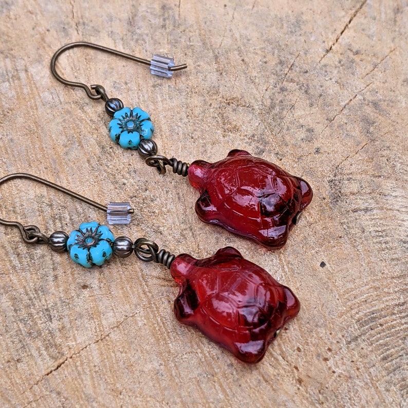 May include: A pair of earrings featuring a red glass turtle charm and a turquoise blue flower bead on each earring.