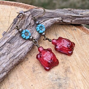 May include: A pair of dangle earrings featuring red glass turtle charms and turquoise blue flower accents. The earrings are made with antique brass wire and beads.