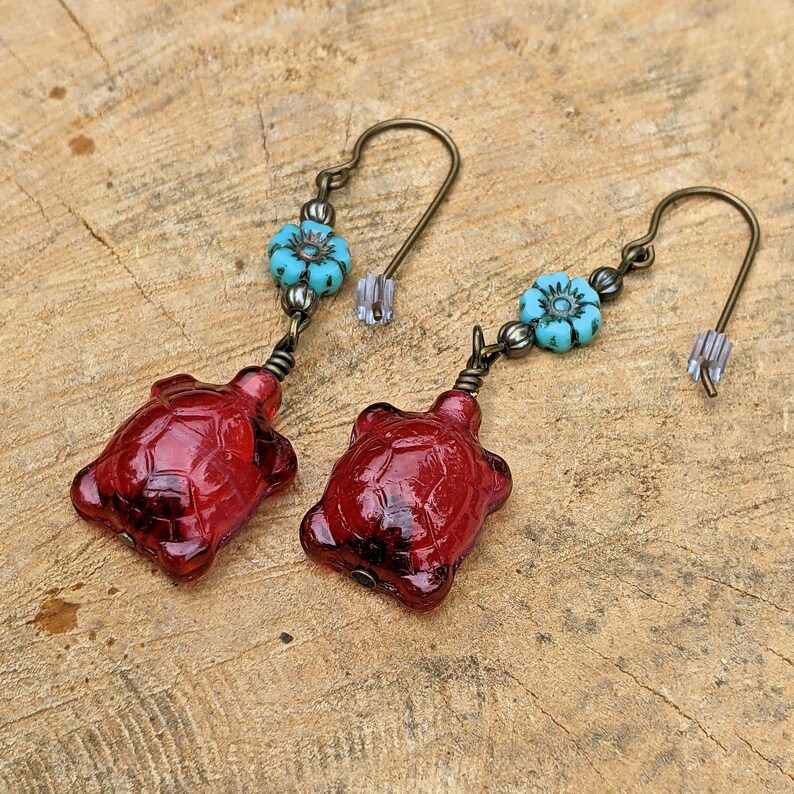 May include: A pair of dangle earrings featuring red glass turtle charms and turquoise blue flower accents. The earrings have a bronze tone.