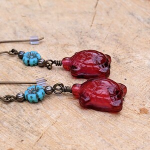 May include: A pair of dangle earrings featuring a red glass turtle charm and a turquoise blue flower bead on each earring.