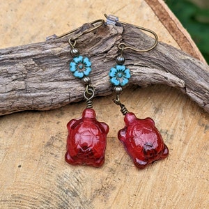 May include: Red glass turtle earrings with turquoise blue flower accents. The earrings are made with bronze wire and beads.