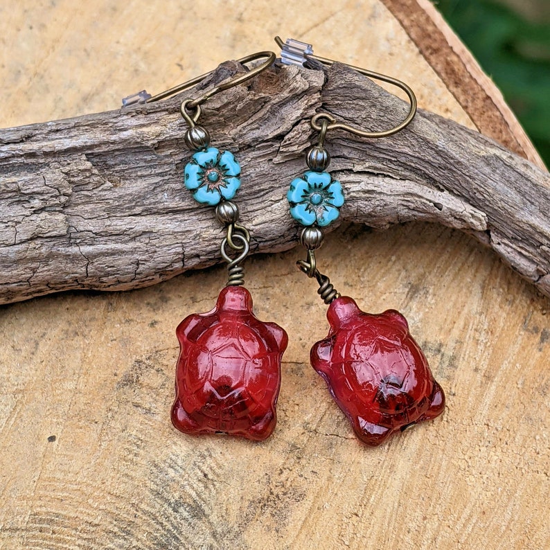 May include: Red glass turtle earrings with turquoise blue flower accents. The earrings are made with bronze wire and beads.