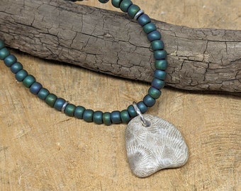 Pode incluir: Um colar com uma corrente de contas verde-escuro iridescente e um pingente de prata feito de pedra natural. O pingente tem uma superfície texturizada única.