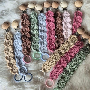 Könnte beinhalten: Eine Auswahl an gehäkelten Schnullerhaltern in verschiedenen Farben wie Beige, Blau, Braun, Grün, Rosa und Mauve. Jeder Halter hat einen Holzclip und einen passenden Ring. Präsentation auf einer weichen, weißen Oberfläche.