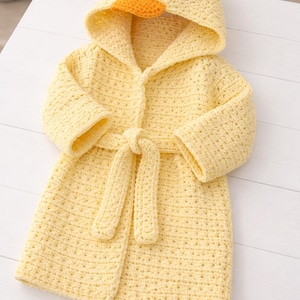 May include: A handmade, yellow crochet bathrobe with a duck hood. The hood features an orange beak and black eyes. The robe has a matching belt and is displayed on a white surface. A cozy, whimsical garment.