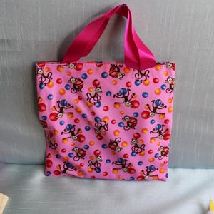 May include: Pink tote bag featuring a pattern of cartoon monkeys and colourful polka dots. The bag has bright pink ribbon handles.