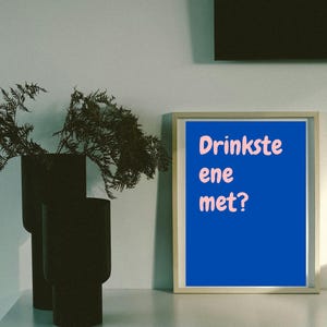 May include: A framed print with the text "Drinkste ene met?" in pink on a blue background. Two black vases with greenery are to the left of the print. The print is in a light-coloured wooden frame.