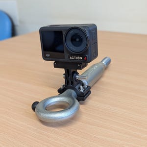 May include: A black action camera labeled "ACTION 4" is mounted on a silver metal tow hook. The camera is attached to a black bracket with screws. The setup is on a light brown wooden surface.