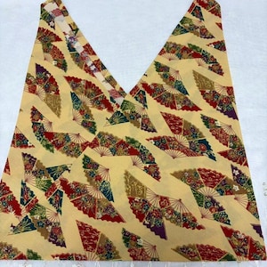 May include: A folded piece of fabric with a repeating pattern of colorful fans on a light yellow background. The fans feature red, green, gold, and purple accents. The fabric is folded into a V-shape, showcasing the pattern's design.