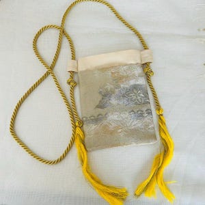 以下が含まれることがあります： 金色のロープストラップと黄色のタッセルが付いた、小さくて長方形のショルダーバッグ。 バッグの本体は、花柄のくすんだ金色です。 バッグの上部はクリーム色です。 バッグは肩にかけるようにデザインされています。