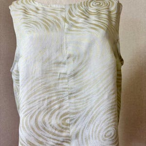 Puede incluir: Una blusa sin mangas con base blanca y un patrón repetitivo de líneas doradas metálicas en espiral. La prenda presenta un cuello redondo y una costura vertical que recorre el centro de la parte delantera, confeccionada en tela fluida.