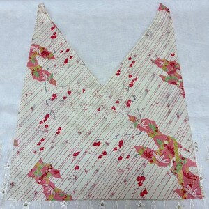 May include: A folded, cream-colored fabric with a diagonal striped pattern in red. The fabric features pink and green floral designs. The fabric is folded into a triangular shape.